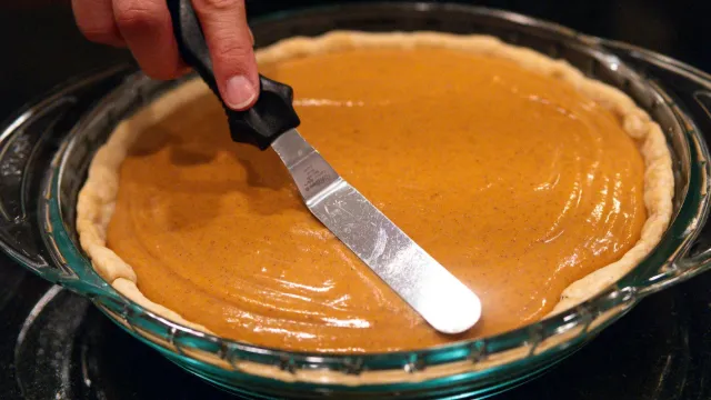 A hand holding an offset spatula smooths unbaked pumpkin pie filling into a pastry crust in a glass pie dish