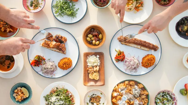 Overhead view of a table spread with more than a dozen shared dishes: grilled kebabs with bulgur and pickled onions, olives, greens, soups, dumplings in yogurt and small salads, with several hands reaching in with forks