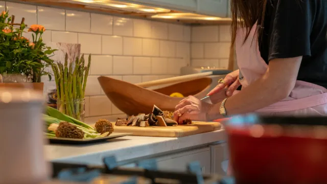 A woman in a pink apron, seen from the shoulders down, chops mushrooms on a wooden board at a warmly lit kitchen counter, with morels and asparagus beside her
